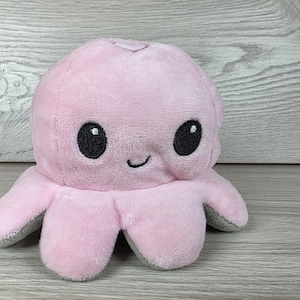 May include: A soft plush octopus toy featuring a light pink exterior with a smooth texture. The octopus has two large, round, black embroidered eyes and a small, curved black embroidered mouth, with grey fabric visible on the underside of its tentacles.