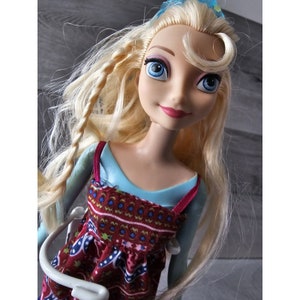 Puede incluir: Una mu&ntilde;eca rubia con ojos azules que lleva un vestido estampado en azul y rojo. La mu&ntilde;eca tiene una trenza en el pelo y lleva una diadema azul con una flor blanca.