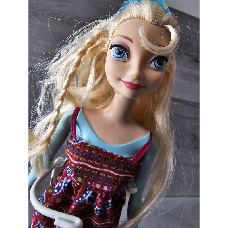 Puede incluir: Una mu&ntilde;eca rubia con ojos azules que lleva un vestido estampado en azul y rojo. La mu&ntilde;eca tiene una trenza en el pelo y lleva una diadema azul con una flor blanca.