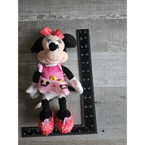 May include: A pink and black plush toy of Minnie Mouse wearing a pink dress with a yellow button and a pink and white skirt. She has a red bow in her hair and is holding a pink and white cupcake.