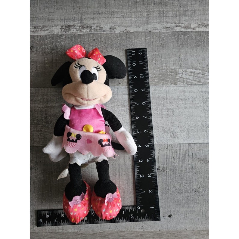 May include: A pink and black plush toy of Minnie Mouse wearing a pink dress with a yellow button and a pink and white skirt. She has a red bow in her hair and is holding a pink and white cupcake.