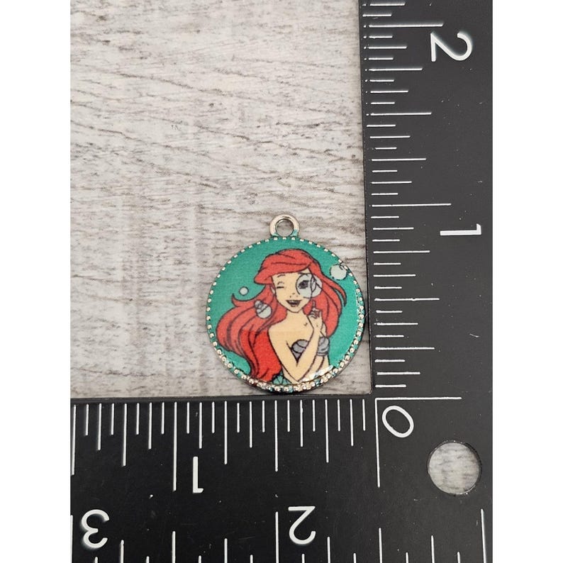 May include: A round, silver charm with a green background and a picture of Ariel from The Little Mermaid. Ariel is wearing a seashell bra and has red hair. The charm is about 1 inch in diameter.