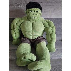 May include: Green plush Hulk toy sitting with a serious expression. The toy has a muscular build and is wearing black shorts.