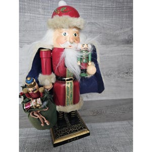May include: A wooden Santa Claus nutcracker holding a smaller nutcracker and a bag of gifts. The Santa Claus nutcracker is wearing a red and white suit with a blue cape. The base of the nutcracker is black with gold accents and the year 2002 is printed on the base.