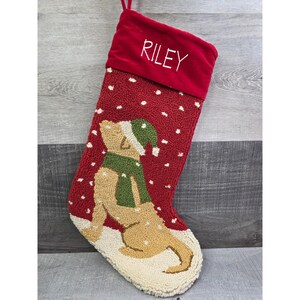Puede incluir: Calcetín navideño rojo con diseño de perro. El calcetín tiene un puño de terciopelo rojo con el nombre "RILEY" bordado en blanco. El cuerpo es rojo con lunares blancos y un perro marrón con bufanda y gorro verdes.