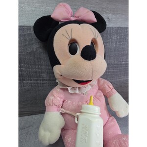 Puede incluir: Mu&ntilde;eca de peluche de Minnie Mouse con un vestido rosa con estampados florales blancos y un lazo a juego. La mu&ntilde;eca sostiene un biber&oacute;n blanco con una tetina amarilla. La mu&ntilde;eca tiene orejas negras y una cara rosa.