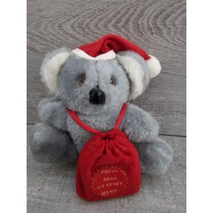 May include: A gray plush koala wearing a red Santa hat and carrying a red bag with the words "Press Here To Start Music" on it.