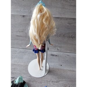 Puede incluir: Una mu&ntilde;eca rubia con una corona azul y blanca en la cabeza lleva un vestido estampado en azul y rojo. La mu&ntilde;eca est&aacute; de pie sobre un soporte de mu&ntilde;eca blanco.