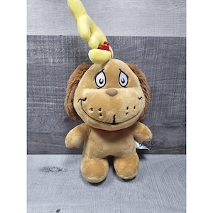 May include: A soft toy dog with brown fur, floppy ears, and a red collar. The dog has a smiling face with large, round eyes. A yellow hand with a red bow is attached to the dog's head.