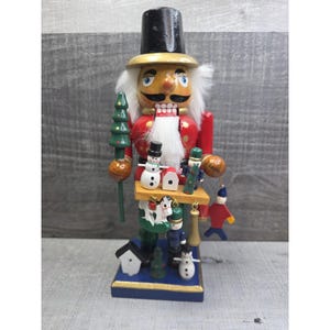 May include: A wooden Christmas nutcracker figurine, featuring a red jacket, black top hat, and white beard. The nutcracker holds a green tree and a small platform with miniature holiday figures, including snowmen and a house. The base is blue with gold trim.