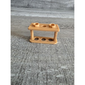 May include: A small, tan-colored test tube rack with two circular holders on top and three holes below. The rack is made of a solid material and has a simple, functional design. Ideal for science-themed decor or educational purposes.