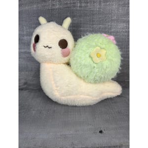 May include: A plush snail toy with a cream-colored body, brown eyes, and pink blush. The snail's shell is light green with a yellow flower and pink accents. The toy has a soft, fuzzy texture.