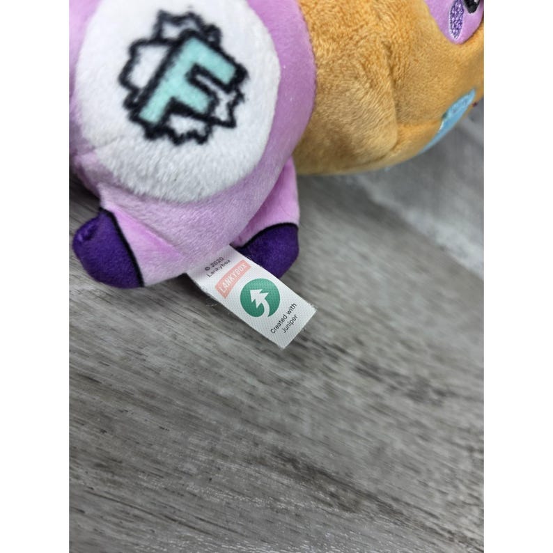 May include: A plush toy featuring a light purple body with a white circular patch on the side displaying a large, stylized mint green letter F. The toy has dark purple accents and a white fabric tag with the text Lankybox and Created with Juniper.