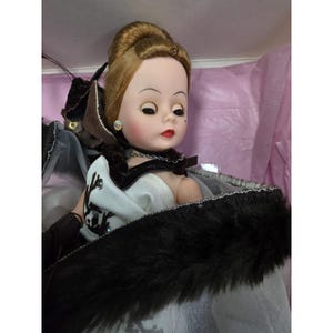 May include: A vintage doll with blonde hair in an updo, wearing a white dress and a black fur-trimmed cape. The doll has a delicate face with red lips, earrings, and a necklace. The doll is in a box with pink tissue paper.