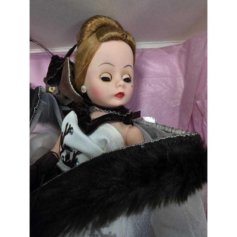 May include: A vintage doll with blonde hair in an updo, wearing a white dress and a black fur-trimmed cape. The doll has a delicate face with red lips, earrings, and a necklace. The doll is in a box with pink tissue paper.