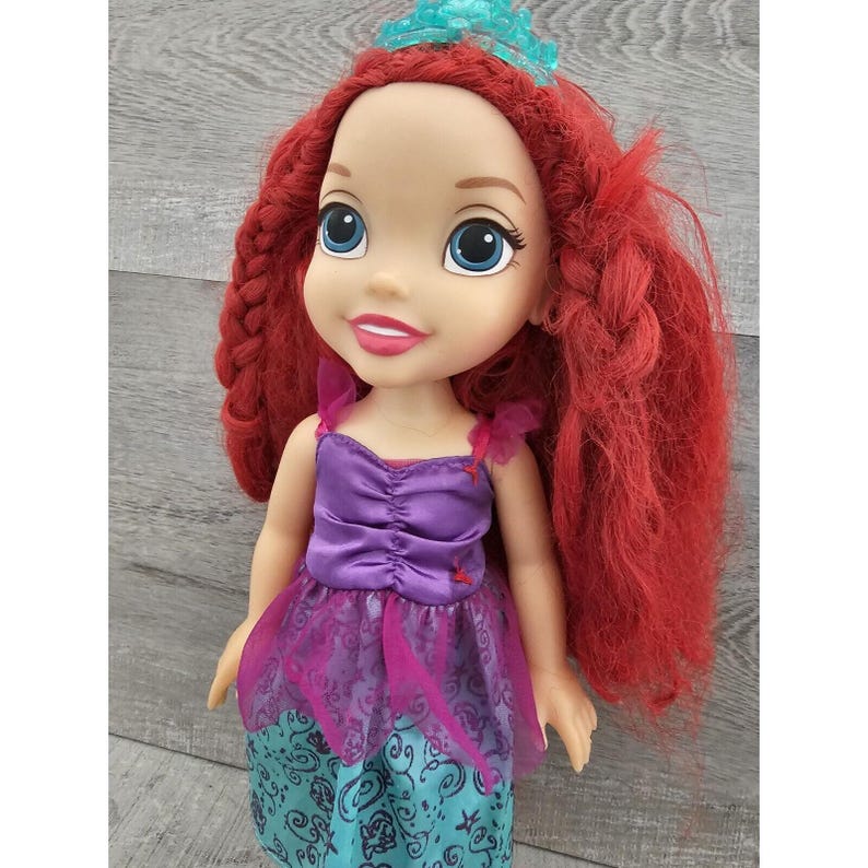 Puede incluir: Una mu&ntilde;eca de pelo rojo con un vestido morado y turquesa con una corona. La mu&ntilde;eca tiene ojos azules y una cara sonriente.