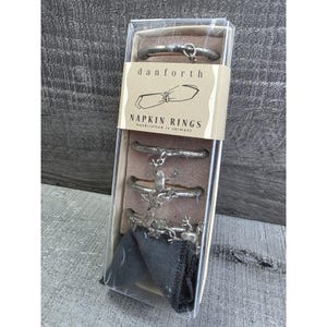 Puede incluir: Un juego de servilleteros Danforth en una caja de plástico transparente. Los anillos son de metal plateado, con una variedad de diseños que incluyen un corazón y ranas. El embalaje incluye un paño negro y una etiqueta que dice "NAPKIN RINGS handcrafted in vermont".