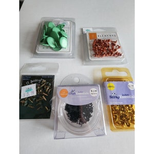 May include: Assortment of craft embellishments in various colors and styles, including green, gold, black, and brown. The embellishments are packaged in clear plastic containers with labels that include the brand name "JoAnn Scrap Essentials" and the price.