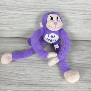 May include: A plush purple monkey toy with long arms and legs, featuring a cream-colored face and belly patch. The belly patch has the text Las Vegas embroidered in purple. The monkey has light-colored hands and feet, and a stitched pink nose.