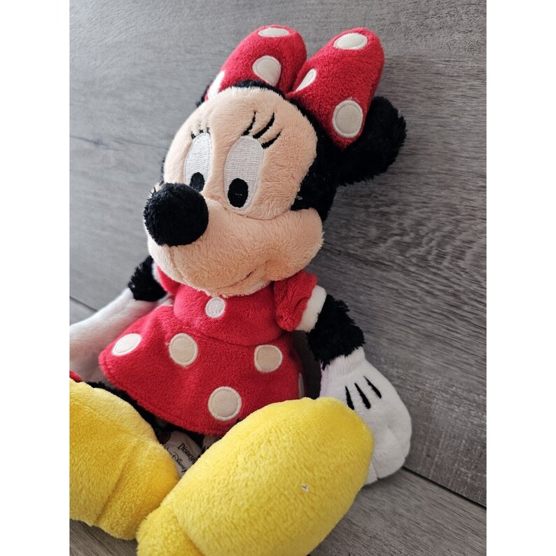 K&ouml;nnte beinhalten: Ein Pl&uuml;schtier von Minnie Mouse, das ein rotes Kleid mit wei&szlig;en Punkten und eine rote Schleife mit wei&szlig;en Punkten tr&auml;gt. Das Spielzeug hat eine schwarze Nase und gro&szlig;e, wei&szlig;e Augen.
