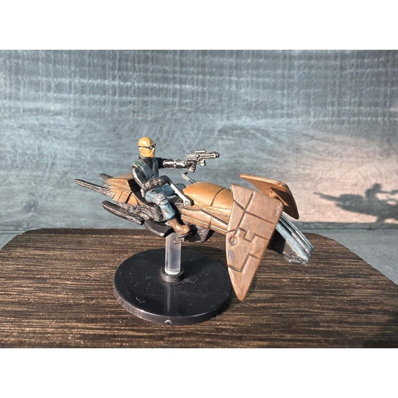 May include: A miniature figurine of a character riding a brown and bronze speeder bike, holding a silver gun. The rider wears a yellow helmet and blue and black clothing. The model is mounted on a black base.