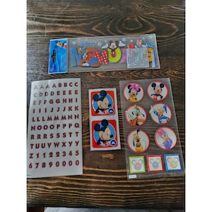 May include: A set of Disney character stickers featuring Mickey Mouse, Donald Duck, Goofy, and Pluto. The stickers are on a clear sheet with a white background. The stickers are perfect for decorating notebooks, planners, and other items.