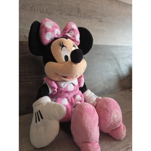 May include: A plush Minnie Mouse doll, wearing a pink dress with white polka dots, a matching bow, and white gloves. The doll has black ears and a black nose. The doll is sitting on a wooden surface.