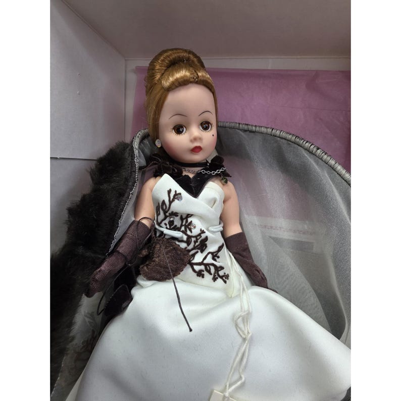 May include: A vintage doll with auburn hair in an updo, wearing a white dress with brown floral embroidery. The doll has brown gloves and a black choker. A dark fur wrap is behind the doll, which is displayed in a box.