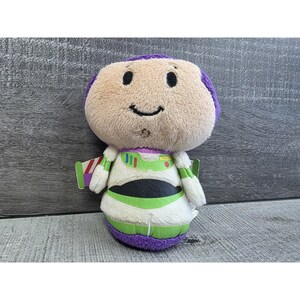 May include: A plush Buzz Lightyear doll from the Toy Story franchise. The doll features a tan face with a purple head, black eyes, and a stitched smile. It wears a white and green spacesuit with purple accents and small wings.