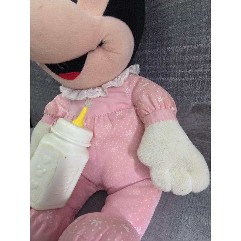 Puede incluir: Mu&ntilde;eca de peluche de Minnie Mouse con un vestido rosa con estampados florales blancos y un cuello de encaje blanco. La mu&ntilde;eca sostiene un biber&oacute;n blanco con una tetina amarilla. La cara de la mu&ntilde;eca es rosa con boca roja y ojos negros.