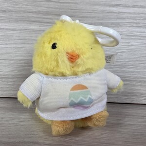 May include: A small, fluffy yellow plush chick keyring featuring a white t-shirt with a graphic print of a pastel-coloured egg with a zigzag pattern. The chick has a small orange beak and black eyes, with a white plastic clip attached to the top.