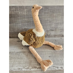 Puede incluir: Un juguete de peluche de avestruz con un cuello y cabeza largos de color beige, cuerpo marrón y patas beige. El juguete tiene detalles blancos alrededor del cuello y en las puntas de las alas. La avestruz está sentada sobre una superficie de madera.