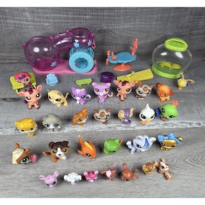 May include: A selection of small plastic animal figurines in various colours, including cats, dogs, and birds. Accessories include a hamster habitat, a small table, and a clear container. The toys are in different poses, suitable for children's play.