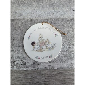 May include: A white ceramic Christmas ornament with a gold string for hanging. The ornament features a Nativity scene with cherubic figures and the text "Prepare Ye the Way of the Lord" and the year "1988".