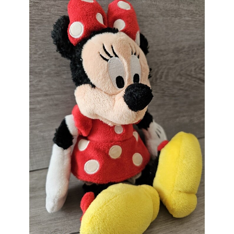 K&ouml;nnte beinhalten: Ein Pl&uuml;schtier von Minnie Mouse, die ein rotes Kleid mit wei&szlig;en Punkten und eine rote Schleife mit wei&szlig;en Punkten tr&auml;gt. Sie hat schwarze Augen, Wimpern und eine schwarze Nase. Ihre Schuhe sind gelb.