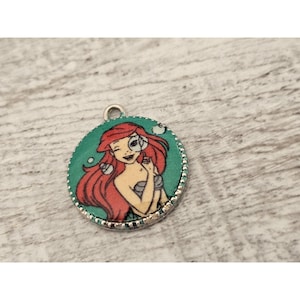 May include: A round silver charm with a green background and a picture of Ariel, the Little Mermaid, with red hair and a seashell in her hand.
