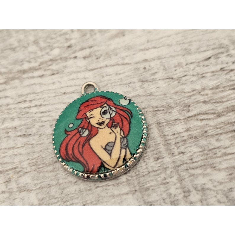 May include: A round silver charm with a green background and a picture of Ariel, the Little Mermaid, with red hair and a seashell in her hand.