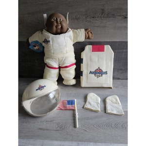 May include: A black Cabbage Patch Kid doll dressed as an astronaut. The doll is wearing a white jumpsuit with red trim and a helmet. The doll is also holding a small American flag. The doll is sitting on a wooden surface.
