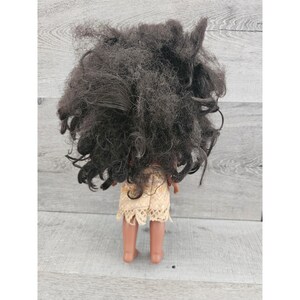 Puede incluir: Una mu&ntilde;eca de piel morena con el pelo largo y negro rizado que lleva un vestido beige y marr&oacute;n con ribete de encaje. La mu&ntilde;eca est&aacute; de pie sobre una superficie de madera.