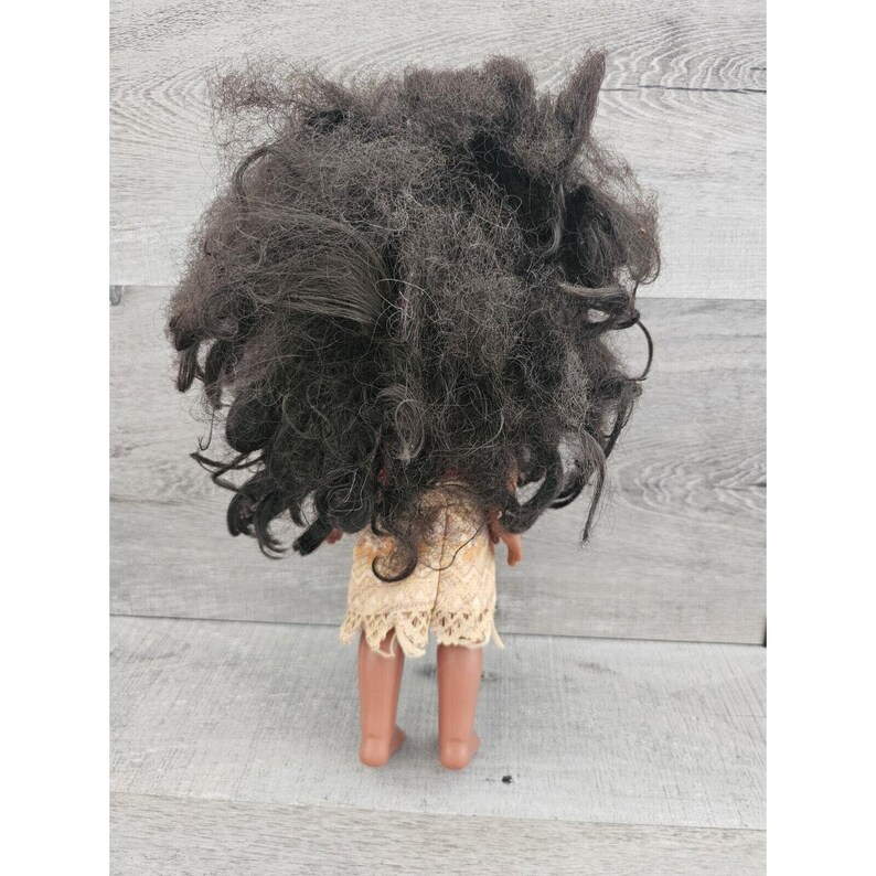 Puede incluir: Una mu&ntilde;eca de piel morena con el pelo largo y negro rizado que lleva un vestido beige y marr&oacute;n con ribete de encaje. La mu&ntilde;eca est&aacute; de pie sobre una superficie de madera.
