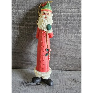 Op de afbeelding: Een lange kerstversiering van de kerstman. De versiering is rood met witte randen en een groene hoed. De kerstman heeft een witte baard en houdt een groene bal vast. Er is een gouden touwtje bevestigd om op te hangen.