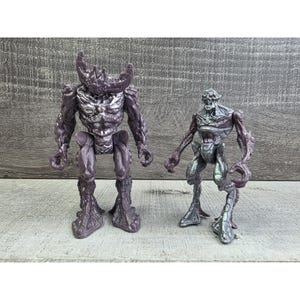 Peut inclure: Deux figurines de monstres violets et argentés. La plus grande figurine a des cornes et une poitrine détaillée. La plus petite figurine a un bras tordu. Les deux figurines ont des textures détaillées.