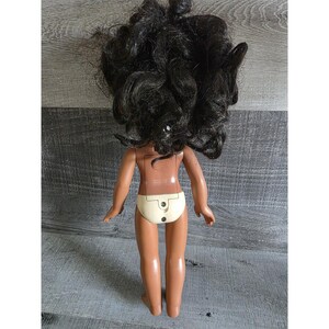 May include: A vintage doll with dark curly hair and tan skin. The doll is wearing a cream-colored diaper. The back of the doll has a battery compartment. The doll's arms are outstretched.