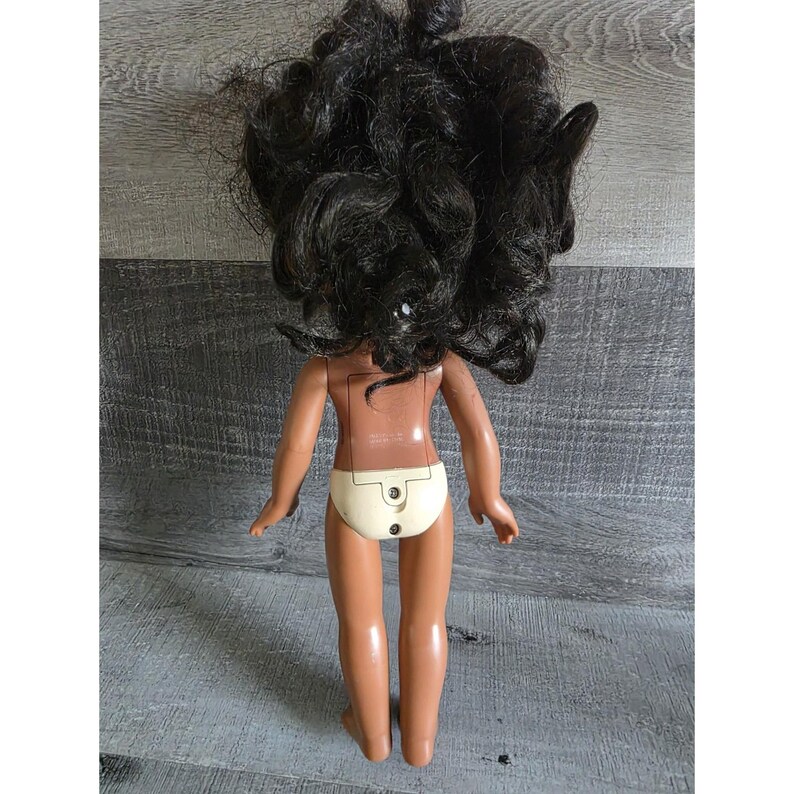 May include: A vintage doll with dark curly hair and tan skin. The doll is wearing a cream-colored diaper. The back of the doll has a battery compartment. The doll's arms are outstretched.