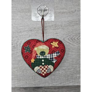 May include: Red felt heart-shaped ornament with a patchwork design featuring a bear wearing a green and white plaid scarf and a red hat. The bear is holding a red and white heart. The ornament has a string for hanging.