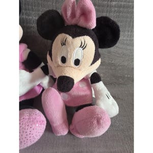 May include: A plush Minnie Mouse doll with a black head, ears, and a pink bow. The doll has a tan face, a pink polka-dot dress, and pink legs. The hands are white with black gloves. The doll is sitting on a gray surface.