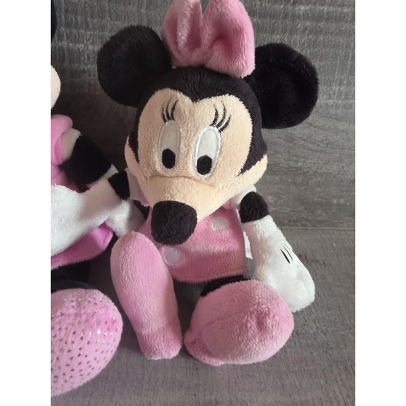 May include: A plush Minnie Mouse doll with a black head, ears, and a pink bow. The doll has a tan face, a pink polka-dot dress, and pink legs. The hands are white with black gloves. The doll is sitting on a gray surface.