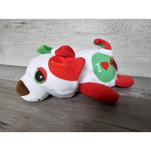 Pode incluir: Brinquedo de pelúcia branco em forma de cachorro com orelhas vermelhas e um remendo verde nas costas. O remendo tem um coração branco com o texto "I" na frente. O cachorro está deitado de lado com a cabeça apoiada no chão.
