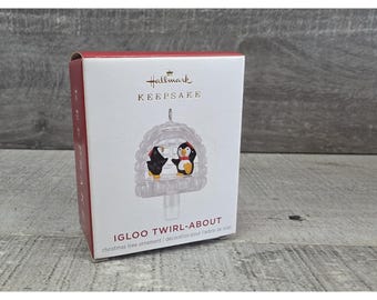 May include: A Hallmark Keepsake Christmas tree ornament box. The box is red and white with the words "Hallmark Keepsake" printed on the front. The ornament is an igloo with two penguins inside. The text "IGLOO TWIRL-ABOUT" is printed on the box.