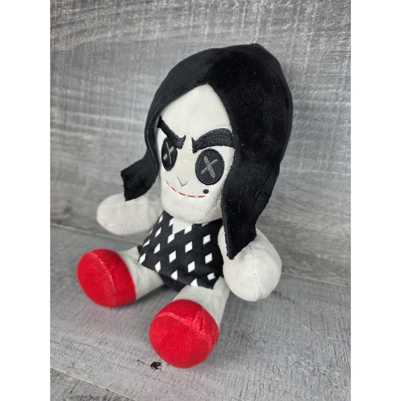 May include: A plush doll with a white face, black hair, and red feet. The doll has button eyes with an X design and a black and white diamond pattern on the dress. The doll is sitting down.